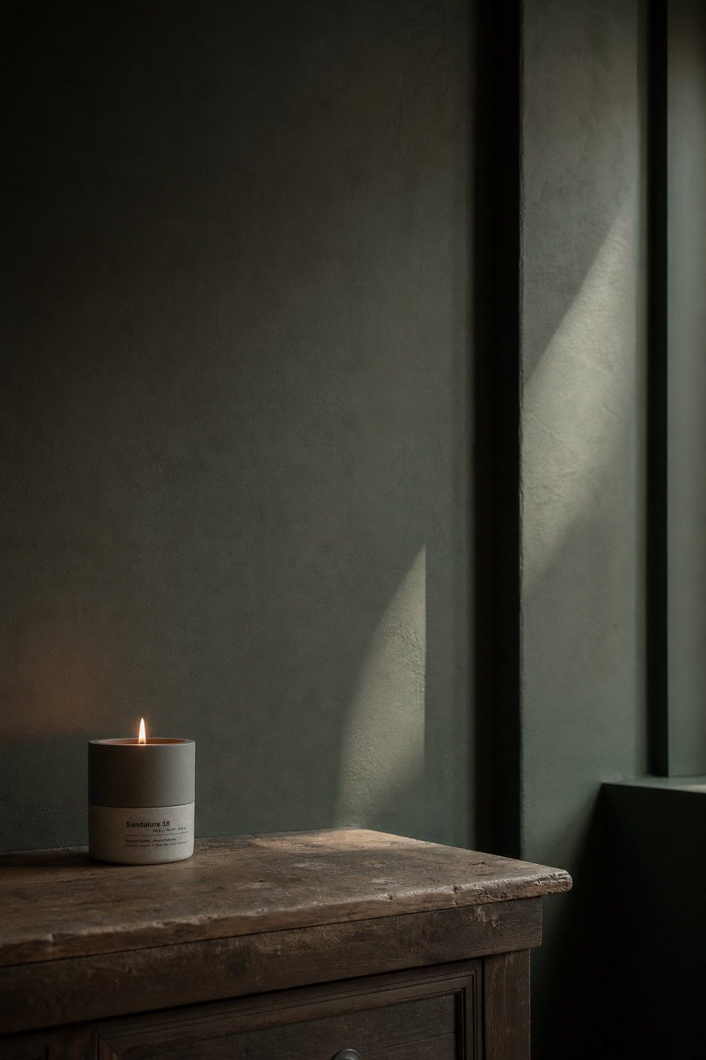 Sandalure 18 concrete soy candle burning on aged wooden dresser in dark green-gray plastered room, diagonal window light on wall — Stāndle