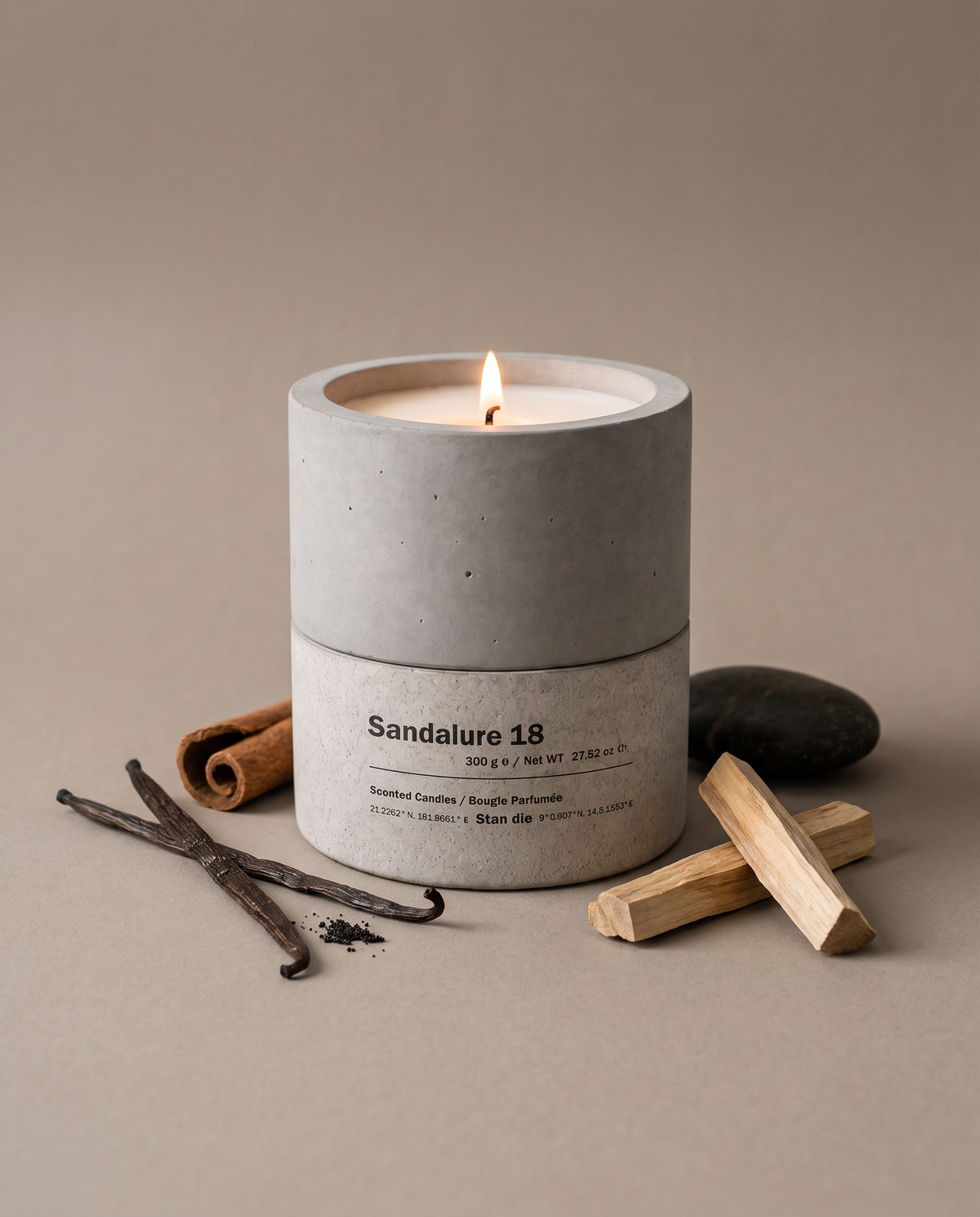 Sandalure 18 sandalwood and vanilla concrete soy candle by Stāndle, lit, beside split vanilla pods, a cinnamon stick, pale sandalwood pieces and a dark river stone.