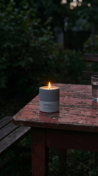 Sandalure 18 concrete candle burning on a weathered red outdoor table at dusk, beside a water glass and dark red grapes on a wooden board, straw hat on chair in foreground