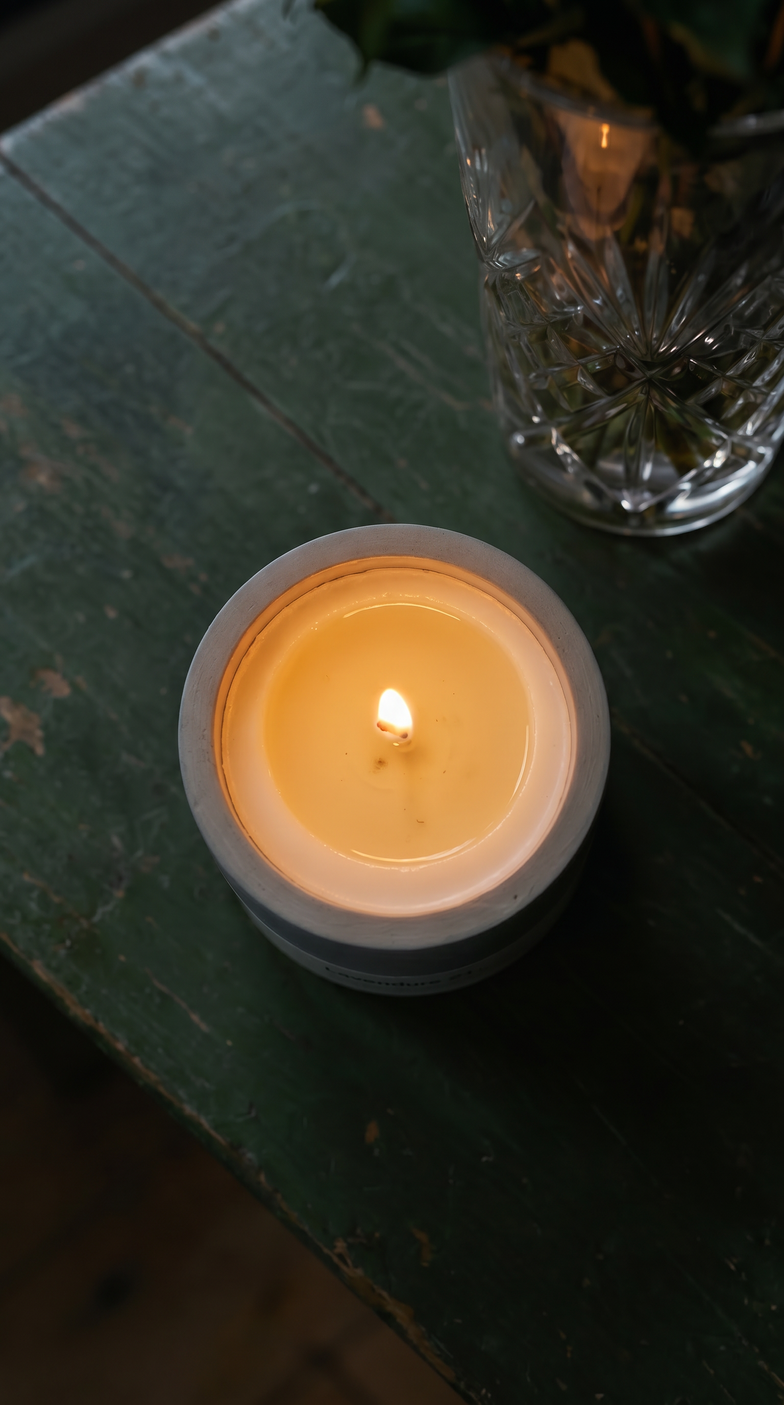 Top-down view of a burning Stān dle concrete candle with glowing amber wax pool, beside a cut crystal vase with greenery on an aged dark green wooden table
