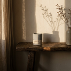 Sandalure 18 sandalwood and vanilla candle on a live-edge wood console in low morning light, beside a linen curtain and a stoneware vase of dried branches